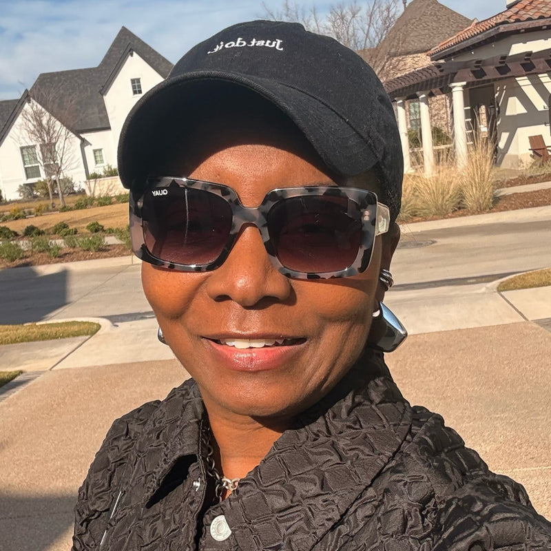 Michelle woman wearing a black baseball cap with white script text, tortoiseshell square sunglasses, and a textured black jacket, photographed outdoors in front of suburban houses.